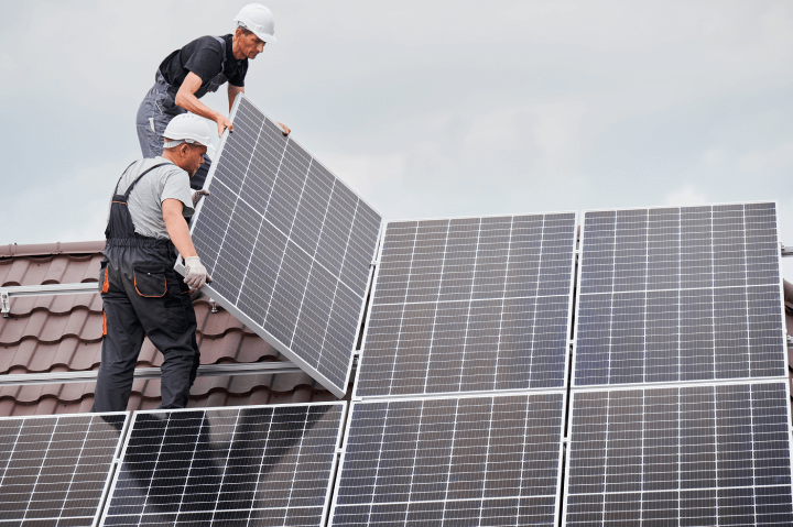 Male technicians are installing photovoltaic solar modules on the roof of a house. Builders wearing helmets are setting up a solar panel system outside, representing the concept of alternative and renewable energy.