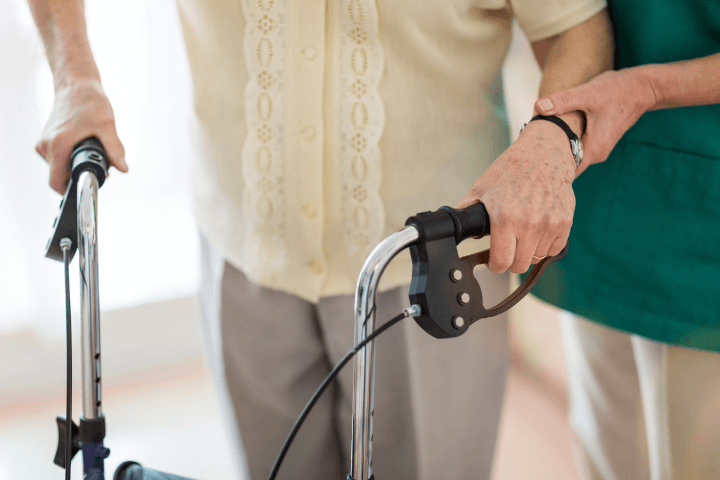 The nursing assistant is assisting an elderly woman by providing support with a walking frame.