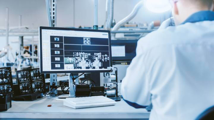 An electronics assembly professional, dressed in a white work coat, examines a printed circuit board displayed on a computer monitor linked to a digital microscope, within a state-of-the-art high-tech factory.