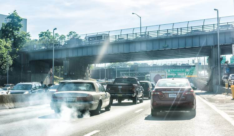 Street and highway in NYC featuring signs for the George Washington Bridge, with smoke emitting from a car's exhaust pipe.