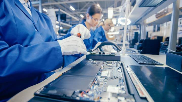 Close-up photograph depicting a female worker in a blue work coat assembling a laptop's motherboard using a screwdriver. The image showcases a high-tech factory facility bustling with multiple employees.