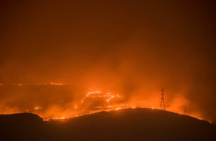 Nighttime view of a large wildfire approaching Grand Coulee, Washington, with the city's lights and fire glow visible through smoky air. Distinct silhouettes of power line towers connecting to the Grand Coulee Dam are evident.