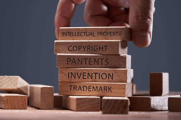 Image of a person placing a block labeled 'Intellectual Property' on top of another wooden block. The blocks also have the text 'Copyright,' 'Patents,' 'Invention,' and 'Trademark'.